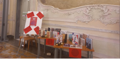 Un allestimento espositivo di libri su tavoli in legno all'interno di un elegante palazzo storico con decorazioni barocche e stucchi ornamentali.