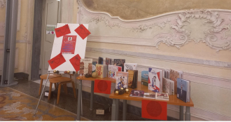 Un allestimento espositivo di libri su tavoli in legno all'interno di un elegante palazzo storico con decorazioni barocche e stucchi ornamentali.